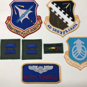 May include: A collection of seven embroidered patches, including a blue and white patch with the text "USAF TEST PILOT SCHOOL", a green and blue patch with the text "AF SYSTEMS COMMAND", a brown and white patch with the text "AD INEXPLORATA", a green patch with the text "AVIATION METAL GOING GOING GONE", and three green patches with blue bars.