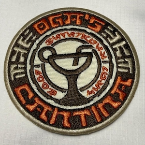 Könnte beinhalten: Ein runder gestickter Aufnäher mit einem dunkelbraunen Cocktailglas-Symbol auf cremefarbenem Hintergrund. Der Rand enthält den Text THE OG'S SET und CANTINA in orangefarbenem und dunkelbraunem Garn mit beigem Kettelrand.