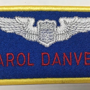Puede incluir: Parche bordado con fondo azul y borde dorado. El parche presenta un emblema blanco con alas y el nombre "CAROL DANVERS" en letras rojas.
