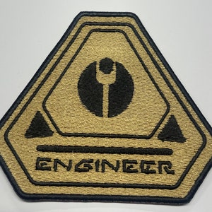 May include: A tan and black embroidered patch with the word "ENGINEER" and a stylized wrench symbol inside a circle.