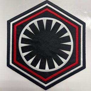 May include: A First Order emblem patch in black, white, and red. The patch is a hexagon shape with a black centre, a white circle with white triangular shapes, and red and white borders. It is likely designed for clothing or accessories.