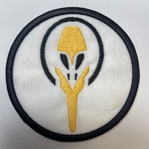 May include: Embroidered patch with a white background, featuring a black outer circle and a black inner circle. The design includes a gold-colored stylized alien head and a gold-colored stem-like shape.