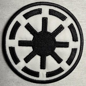 Può includere: Una toppa ricamata circolare con un design in bianco e nero. Il centro è costituito da un cerchio nero pieno con otto raggi. Attorno si trova un anello bianco racchiuso da un bordo nero con otto segmenti rettangolari neri.