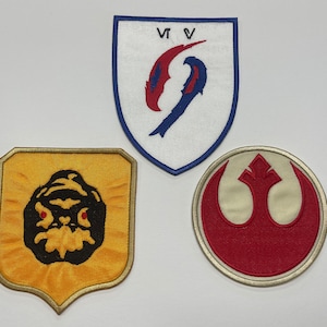 May include: Three embroidered patches. The first patch is white with a blue and red symbol and the text "VVV". The second patch is yellow with a black and red creature. The third patch is cream with a red symbol and a white background.