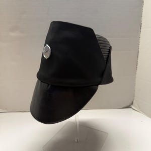 Könnte beinhalten: Schwarzer Hut mit silbernem Emblem auf der Vorderseite. Der Hut hat einen gebogenen Schirm und ein strukturiertes Design. Der Hut wird auf einem transparenten Ständer präsentiert.