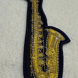 May include: Embroidered patch featuring a gold saxophone on a black background with a navy blue border. The saxophone is intricately detailed with gold thread, showcasing the instrument's keys and curves. A decorative item for music lovers.