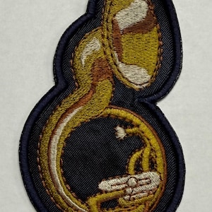 May include: Embroidered patch featuring a sousaphone, a brass instrument. The patch has a navy blue border and a detailed design in shades of gold, brown, and white. The sousaphone is the central focus, showcasing its curved shape and intricate details.