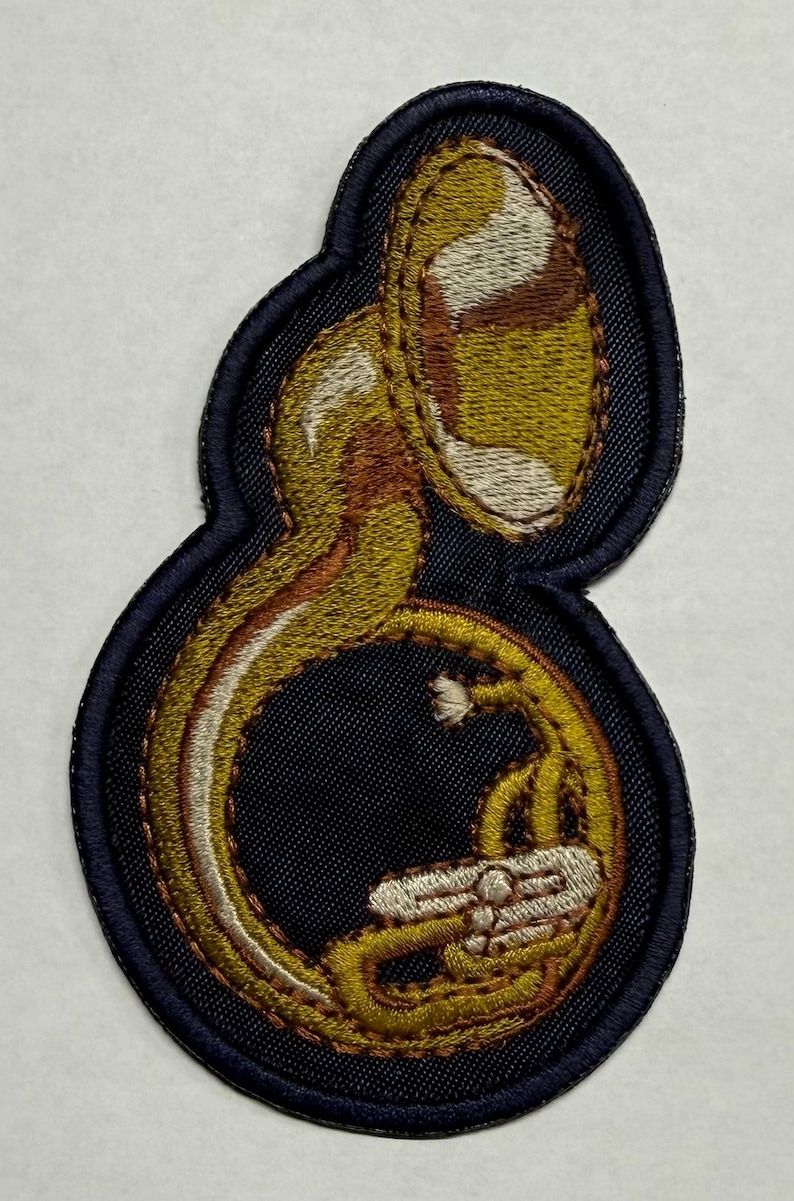 Peut inclure: &Eacute;cusson brod&eacute; repr&eacute;sentant un sousaphone, un instrument de musique en laiton. Le patch a une bordure bleu marine et un motif d&eacute;taill&eacute; dans les tons or, marron et blanc. Le sousaphone est au centre, mettant en valeur sa forme incurv&eacute;e et ses d&eacute;tails.