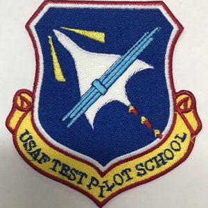 Puede incluir: Parche bordado con un escudo azul, que presenta un avión blanco estilizado y un instrumento azul claro. El parche tiene detalles amarillos y rojos, con el texto "USAF TEST PILOT SCHOOL" en amarillo.