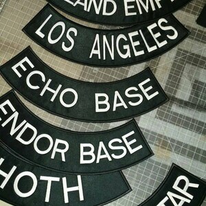 May include: A set of six black and white embroidered patches with curved edges. The patches feature the text "Inland Empire", "Los Angeles", "Echo Base", "Endor Base", "Hoth", and "Death Star".