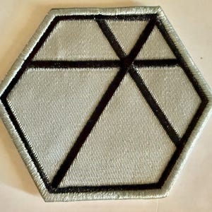Puede incluir: Un parche bordado hexagonal con una base de color gris plateado y una serie de líneas negras que se cruzan creando formas geométricas en la superficie. Los bordes del hexágono están rematados con un borde grueso cosido en gris plateado.