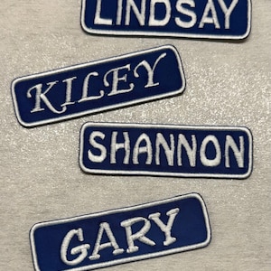 Könnte beinhalten: Vier bestickte Namensaufnäher. Jeder Aufnäher ist ein marineblaues Rechteck mit weißem Rand und weißem gesticktem Text. Die Namen sind Lindsay, Kiley, Shannon und Gary. Geeignet für Kleidung oder Accessoires.