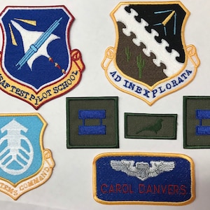 May include: A collection of seven embroidered patches featuring various military insignia. The patches are in different colors and shapes, including blue, green, yellow, and white. The patches feature text such as "USAF Test Pilot School", "AF Systems Command", and "Ad Inexplorata".