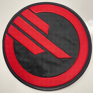 May include: A red and black embroidered patch with a stylized symbol of a red 'S' inside a red circle. The symbol is a popular design from the Star Wars franchise.