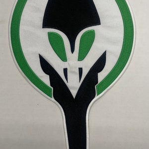 May include: Embroidered patch featuring a stylized design. The patch has a white background with a green border. The central design is a black shape with green accents, resembling a stylized insect or alien head.