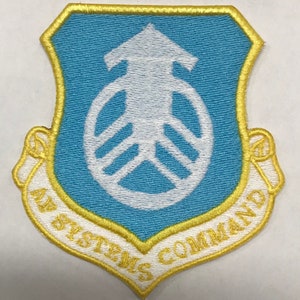 May include: A blue and white embroidered patch with a yellow border. The patch features a white stylized symbol with an arrow pointing upwards. The text "Air Systems Command" is written in a white arc around the symbol.