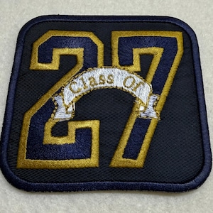 May include: A navy blue embroidered patch with the number "27" in gold and navy blue. A white banner with the words "Class Of" arches across the center. The patch has a navy blue border.