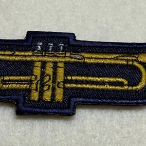 Könnte beinhalten: Gestickter Aufnäher mit einem goldenen Trompeten-Design auf marineblauem Hintergrund. Der Aufnäher hat eine detaillierte Kontur und ist wahrscheinlich für das Aufnähen auf Kleidung oder Accessoires gedacht. Die Trompete ist goldfarben.