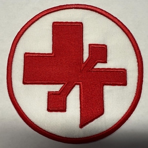May include: A circular patch featuring a red cross-like design within a white circle, bordered by a red stitched edge. The design has a unique, stylized appearance, suitable for apparel or accessories.