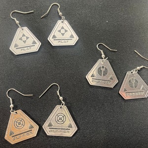 May include: Five silver earrings with triangular pendants. Each pendant has a different design and text. The designs include a star, a wrench, a circle, and two different versions of a target. The text on the pendants reads "Pilot", "Engineer", and "Gunner".