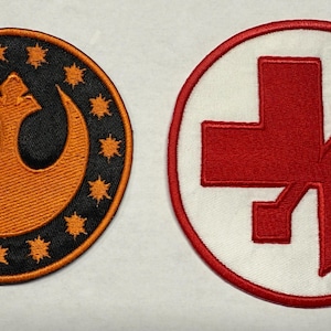 Könnte beinhalten: Zwei gestickte Aufnäher. Einer ist ein schwarzer Kreis mit einem orangefarbenen Emblem und orangenen Sternen. Der andere ist ein weißer Kreis mit einem roten kreuzartigen Symbol. Beide Aufnäher befinden sich auf weißem Hintergrund.