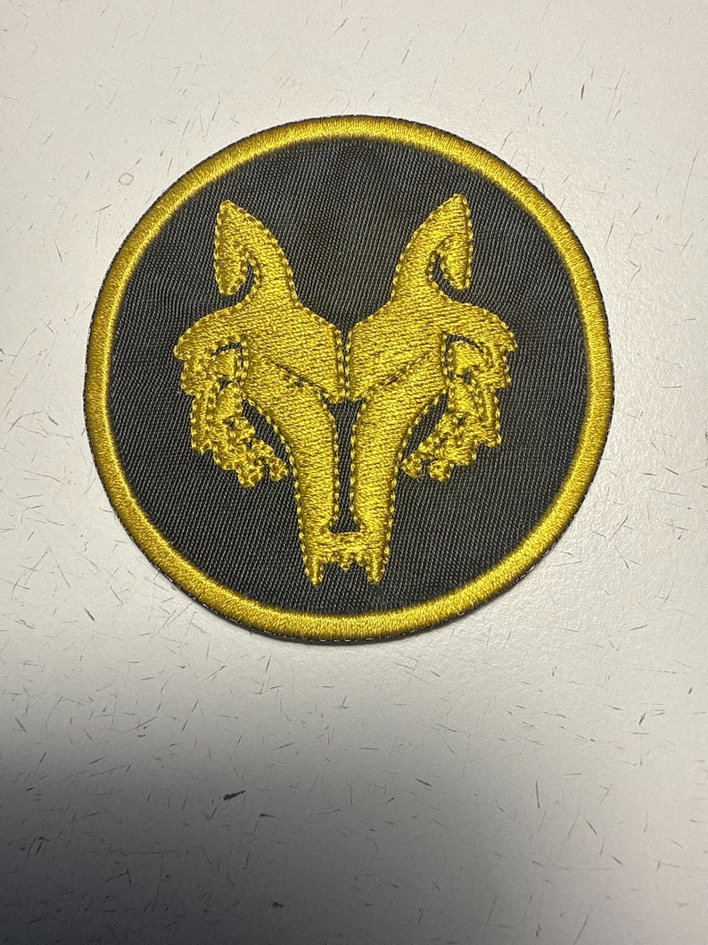 May include: Embroidered patch featuring a symmetrical gold wolf design on a black circular background. The wolf design is detailed with intricate stitching, and the patch has a gold border. This patch is suitable for jackets, bags, or other fabric items.