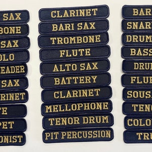 Puede incluir: Un conjunto de parches bordados en azul marino con letras doradas. Los parches presentan varios nombres de instrumentos musicales y roles, como "Tenor Sax," "Clarinete" y "Drum Major." Estos parches son probablemente para uniformes de banda.