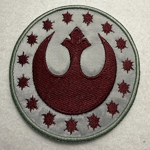 May include: Circular patch featuring a burgundy emblem within a silver field, surrounded by burgundy starbursts and a green border. The emblem is a stylized crescent and crown design. The patch is likely for apparel or accessories.