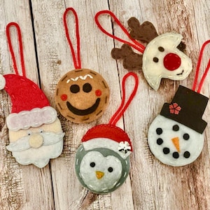 May include: A collection of handmade Christmas ornaments. The ornaments include a Santa Claus, a gingerbread man, a reindeer, a snowman, and a penguin, all made of felt. Each ornament has a red string for hanging.