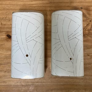 May include: Two semi-cylindrical wall light covers featuring a white surface with a black dashed line pattern of intersecting curved arcs. Each cover has a small circular hole positioned in the lower center section for mounting or hardware attachment.