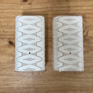 May include: Two semi-cylindrical glass light fixture covers featuring a white geometric diamond pattern on a clear background. Each piece has a small circular hole centered on the front surface, with vertical textured lines along the edges.