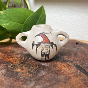 May include: A small, light-colored ceramic vessel with two side handles. The front features a painted graphic of a bird in red and black ink, with a stylized wing design extending downward. The surface has a matte, off-white finish.
