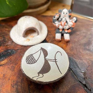 May include: A collection of three ceramic items: a small round bowl with a black line drawing of a figure carrying a basket, a white domed structure with an arched opening, and a small figurine depicting a figure holding three smaller figures.