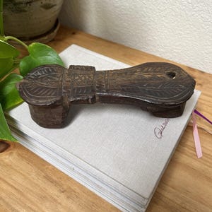 May include: A dark brown, hand-carved wooden object with a flat, elongated shape and a raised, block-like base at one end. The surface features intricate, incised geometric patterns and linear carvings, with a small circular hole near the rounded edge.