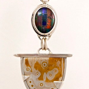 May include: A silver necklace with a pendant featuring a circular, multi-colored stone and a lower, teardrop-shaped stone with a gold and silver pattern.