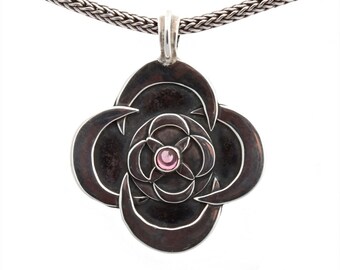 May include: A silver pendant necklace featuring a dark, stylized floral design with a central pink gemstone. The pendant is suspended from a woven silver chain, creating a striking contrast against the white background.