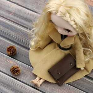 May include: A doll with blonde hair wearing a tan coat and holding a brown leather clutch. The doll is lying on a wooden surface, with two small pine cones nearby. The doll's coat has a black belt.
