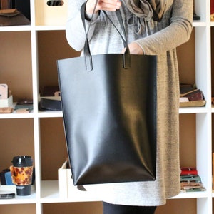 May include: A black leather tote bag with a rectangular shape and two top handles. The bag is held by a person in front of a white shelf with various leather goods.