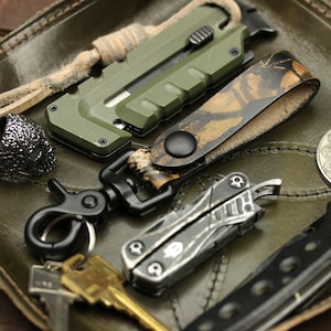 May include: An assortment of everyday carry items, including a green utility knife, a camo leather key holder, a multi-tool, a skull ring, a coin with a yin-yang symbol, and keys, all arranged on a green leather tray.