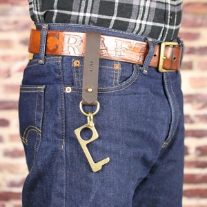 Könnte beinhalten: Ein brauner Ledergürtel mit dem geprägten Wort "CRAFT". Ein brauner Leder-Schlüsselanhänger mit einem Messing-Schlüsselring und einem berührungslosen Werkzeug aus Messing ist an der Gürtelschlaufe befestigt. Der Schlüsselanhänger trägt die Initialen "CAI". Die Person trägt eine blaue Jeans.