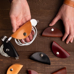 Puede incluir: Surtido de nueve organizadores de cables de cuero en varios colores, incluyendo marrón, negro, rojo y tostado. Los organizadores tienen forma de cáscara de taco y están diseñados para sujetar y organizar cables y cables.