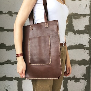 May include: A brown leather tote bag with a front pocket and a stitched design. The bag has two brown leather straps.