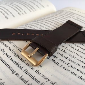 May include: A brown leather watch strap with a gold buckle. The strap is lying on a book with text.