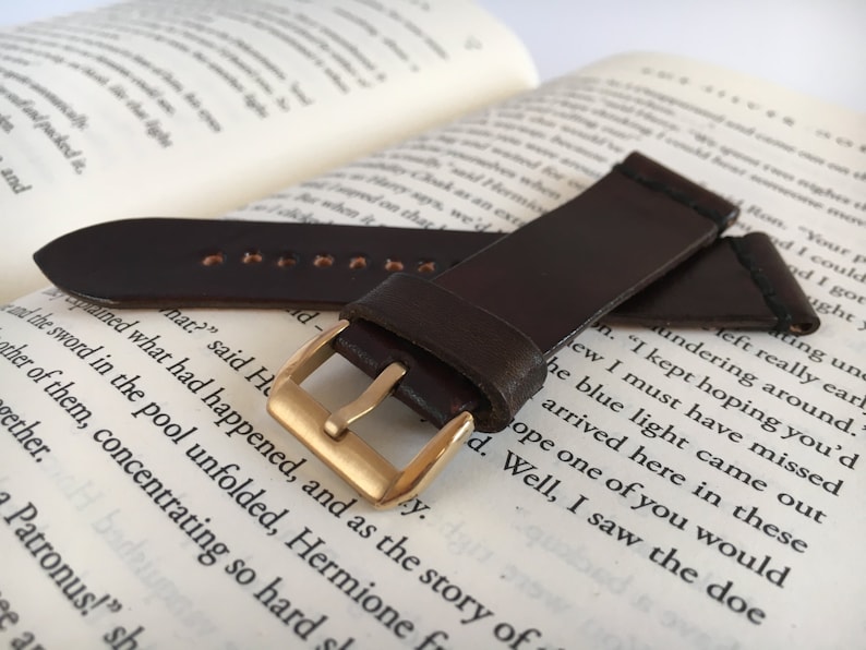 May include: A brown leather watch strap with a gold buckle. The strap is lying on a book with text.
