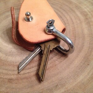 K&ouml;nnte beinhalten: Ein Schl&uuml;sselanh&auml;nger aus Leder mit einem Metallhaken und zwei daran befestigten Schl&uuml;sseln. Der Schl&uuml;sselanh&auml;nger ist hellbraun und hat einen kleinen Metallknopf auf der Vorderseite.
