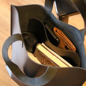 May include: A black leather tote bag with a curved handle, filled with a notebook, wallet, phone, and other items. The bag is open, revealing the contents inside. The interior is a lighter color than the exterior.