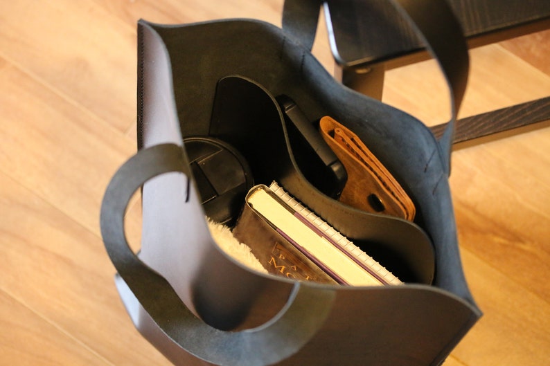 May include: A black leather tote bag with a curved handle, filled with a notebook, wallet, phone, and other items. The bag is open, revealing the contents inside. The interior is a lighter color than the exterior.