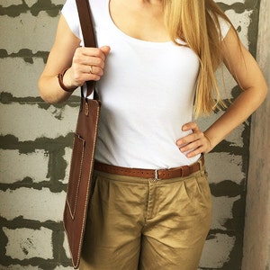 May include: A brown leather tote bag with a front pocket and a long strap. The bag is worn by a person wearing a white t-shirt and khaki pants.