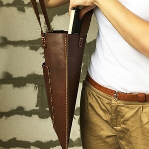 May include: A brown leather tote bag with a front pocket and a long strap. The bag is worn over the shoulder of a person wearing a white shirt and khaki pants.