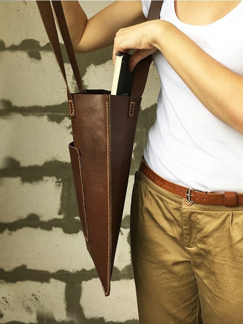 May include: A brown leather tote bag with a front pocket and a long strap. The bag is worn over the shoulder of a person wearing a white shirt and khaki pants.